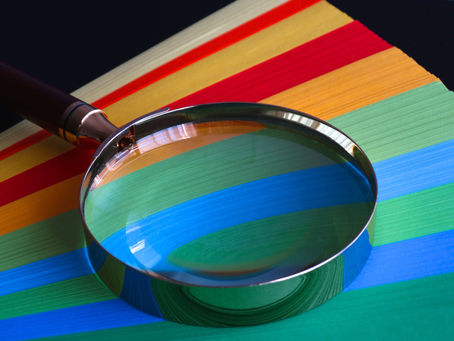 Photograph showing a magnifying glass placed over a stack of colorful paper sheets arranged in horizontal stripes of red, orange, yellow, green, and blue. Magnifying glass enlarges the blue and green stripes, emphasizing texture and details of paper fibers. Testing packaging changes.