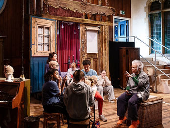 A company of performers sit on the set of a show listening to director Emma Rice