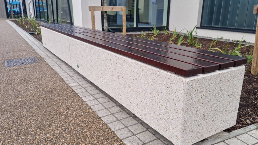 Kube Range Street Seating. A long bench with a polished dark brown wooden top sits on a speckled concrete base. It is alongside a path beside a building.