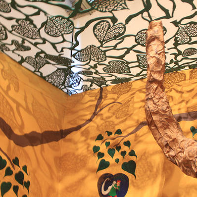 Image of the interior of the exhibit. Wall is painted yellow, a brown paper branch leads to the laser cut peepal leaves roof casting shadow on the yellow wall. The wall is also painted with green leaves showcasing cultural stories around peepal tree.