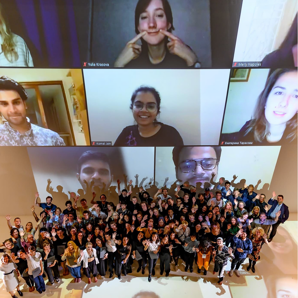The top half is a large projection screen with zoom call. There are eight people on the call. In front of the projection screen are around 100 people cheering and posing for the photo