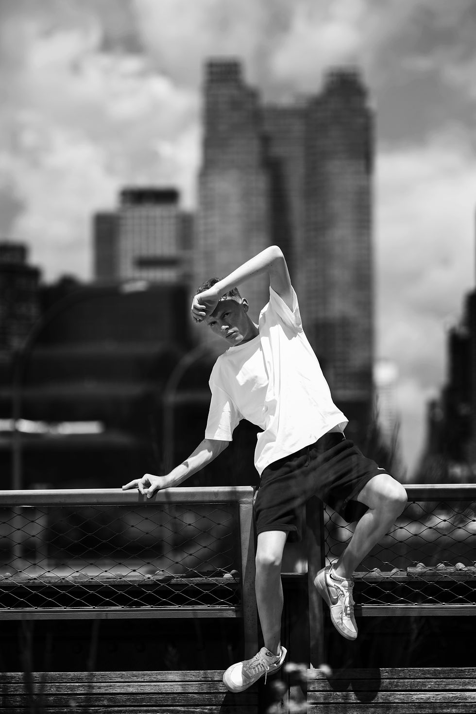Black-and-white photo of model posing on a bridge railing, white T-shirt and black shorts, with blurred city skyscrapers behind him.