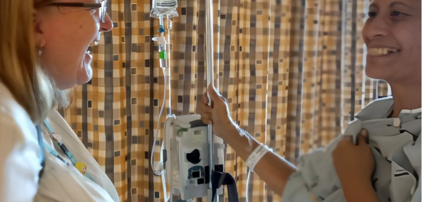 A patient and hospital chaplain stood smiling to each other in front of a hospital curtain
