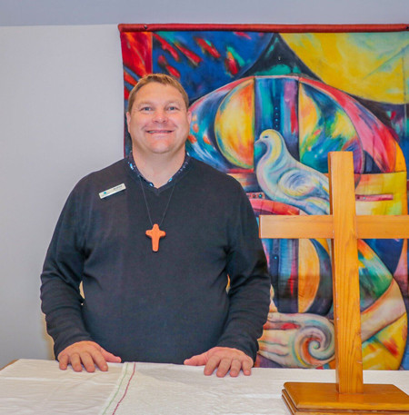 A hospital chaplain smiling next to a wooden cross