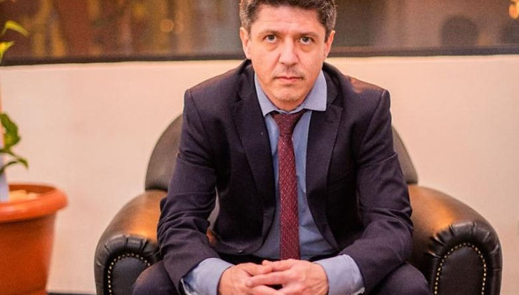 Homem sério, sentado em uma poltrona preta, vestindo um terno escuro, camisa azul clara e gravata vermelha, com as mãos cruzadas sobre os joelhos. Ao fundo, uma parede de vidro reflete luzes pendentes e algumas plantas internas.