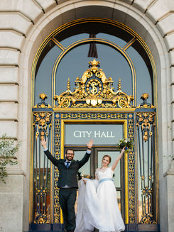 Newlyweds cheering with raised arms at the gold-trimmed entrance of San Francisco City Hall
