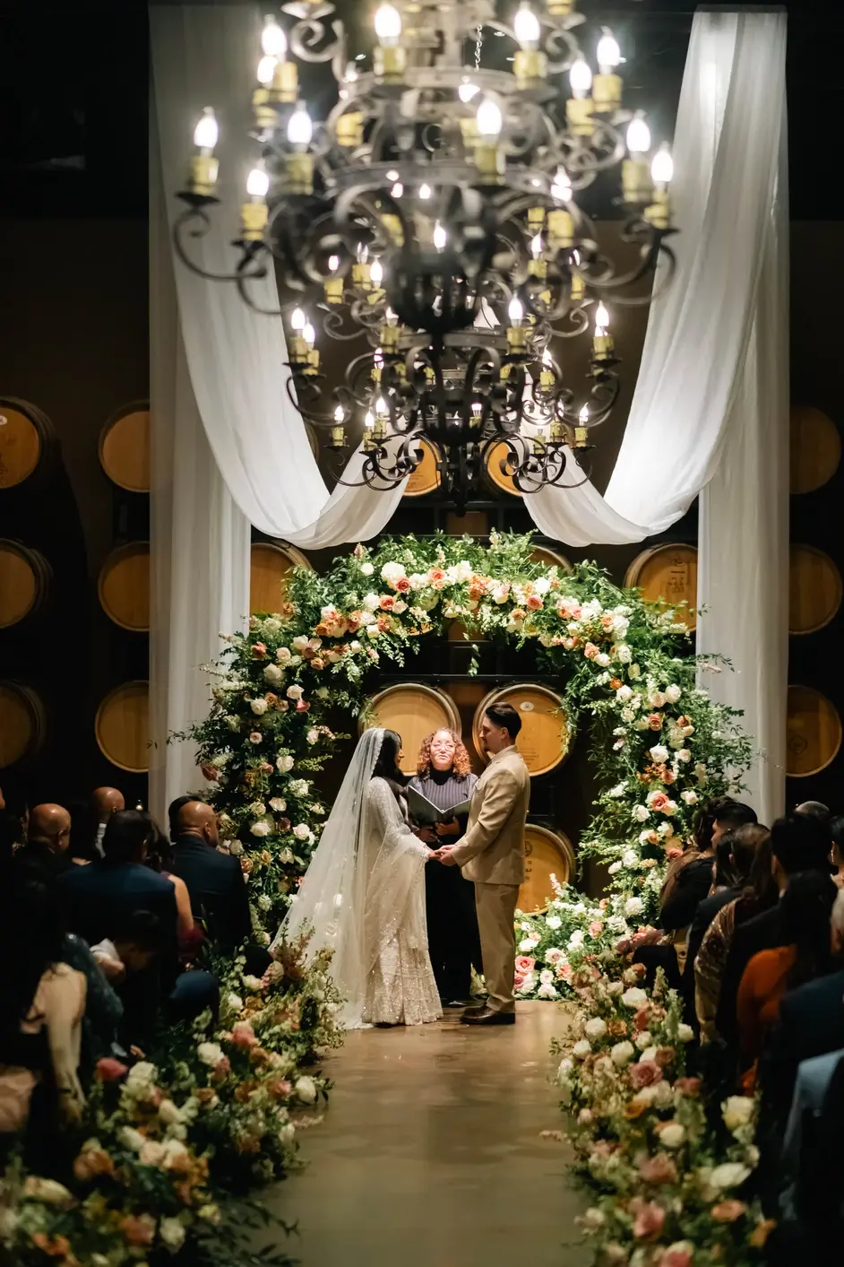 Saundra officiating beneath a floral arch and chandelier as the couple holds hands, wine barrels lining the room behind them