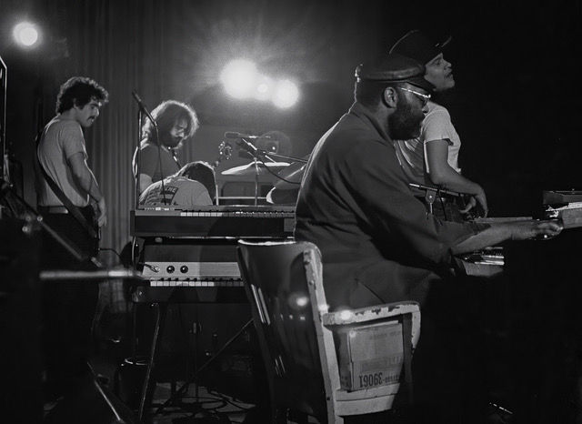 Black and white concert photo of the Jerry Garcia Band performing live on stage, featuring musicians at keyboards and guitar under bright stage lights, circa 1974.