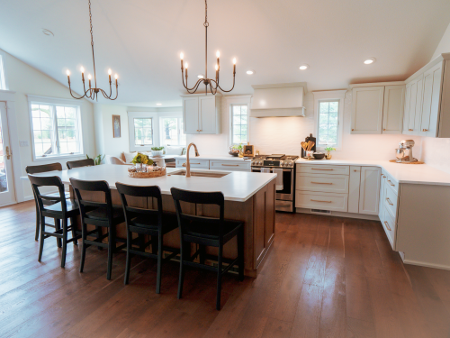 Bright modern white kitchen with large island, black barstools, hanging chandeliers, and stainless stove on wood floors.