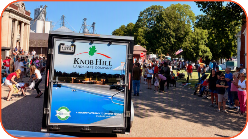 Landscape company trailer with Knob Hill Landscape Company logo rolls past a crowd on a sunny street at a parade.