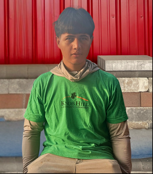 Young man in a green Knob Hill Landscape Company T-shirt sits against a red wall, looking serious.