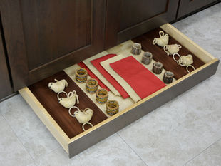Drawer with red and beige napkins, beaded napkin rings, and bird-shaped napkin holders. Set against dark wood and tiled floor. Offered by Dura Supreme Cabinetry at Capitol Kitchen and Bath.