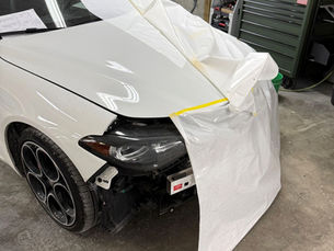 White car in a repair shop with front end masked by paper and tape, exposing the headlight and wheel on a concrete floor.