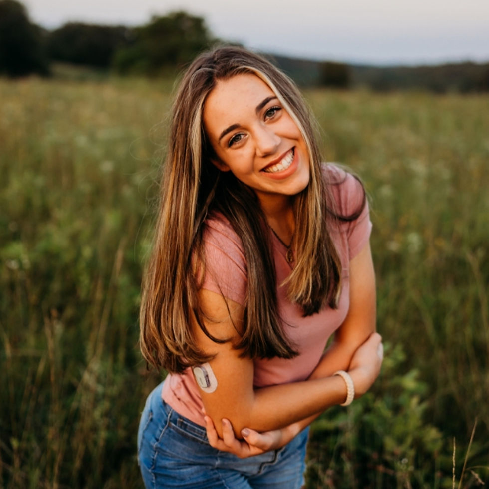 Smiling woman in a pink shirt stands in a grassy field at sunset, a medical sensor visible on her arm, exuding a cheerful mood.