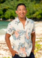 Smiling man in a floral shirt on a beach, hands in pockets. Lush greenery and ocean in background, creating a tropical vibe.