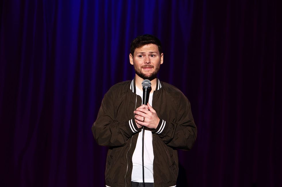 Drew Lynch holding a microphone on stage with a dark purple curtain backdrop. He appears thoughtful, wearing a brown jacket and white shirt.