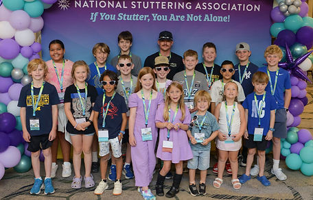 A large group of smiling kids and a few adults pose under a purple and teal balloon arch at an NSA event, with a banner behind them that says 'If You Stutter, You Are Not Alone!