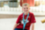 Boy in a red Kansas City Chiefs shirt smiles, wearing a blue lanyard. Background has railings and soft lighting, creating a cheerful mood.