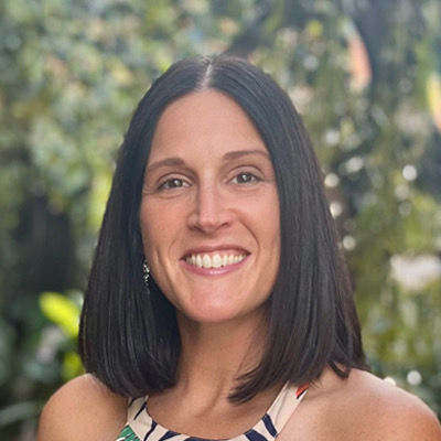 A smiling woman with shoulder-length dark hair in a patterned top stands outdoors, with lush greenery in the blurred background.
