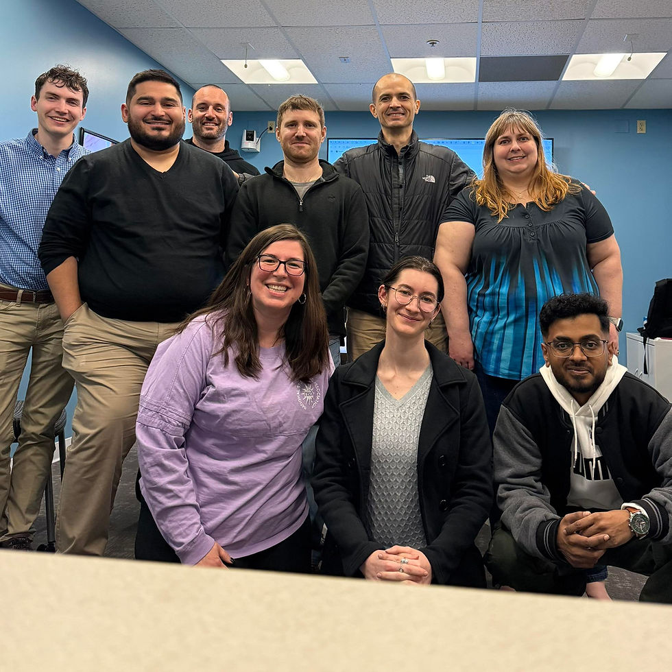 Group of ten people smiling in an office setting with blue walls. They are casually dressed and appear happy. Fluorescent lights above.