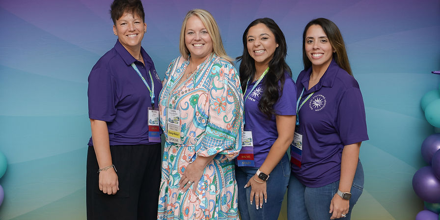 Four smiling women standing indoors in front of a colorful balloon arch and a purple-to-blue gradient backdrop with the National Stuttering Association logo and the phrase ‘If You Stutter, You Are Not Alone!’ Two women wear purple NSA staff shirts, one wears a patterned dress, and one wears a purple NSA crew t-shirt, all with conference badges.