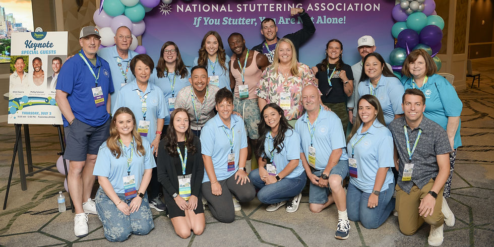 A large group of around 20 people, mostly wearing light blue National Stuttering Association shirts with conference badges, posing cheerfully indoors in front of a balloon arch and a purple backdrop that reads ‘National Stuttering Association: If You Stutter, You Are Not Alone!’ The group includes men and women of diverse backgrounds, smiling and standing or kneeling closely together.