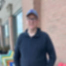 Man in a blue jacket and cap smiles outside a brick building. Colorful chairs line the sidewalk. Relaxed and casual atmosphere.