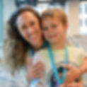 A smiling mother embracing her young son who is wearing a lanyard, both dressed in casual outfits inside a modern building with large windows in the background.