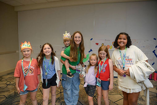 A group of happy kids and a young adult mentor at an NSA event, smiling together in front of a wall filled with colorful artwork.