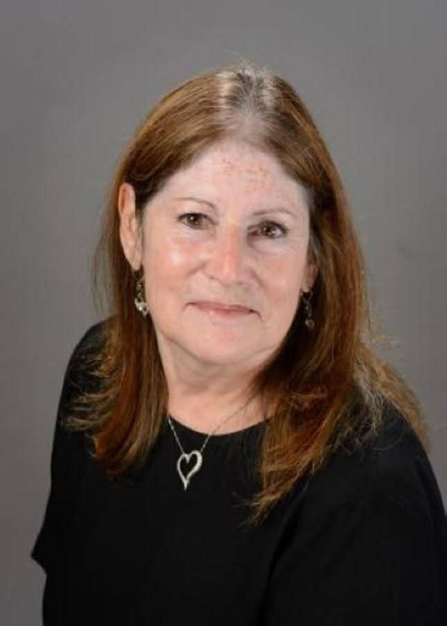 Woman with brown hair, wearing a black top and heart necklace, smiling slightly against a gray background. Mood is calm and neutral.