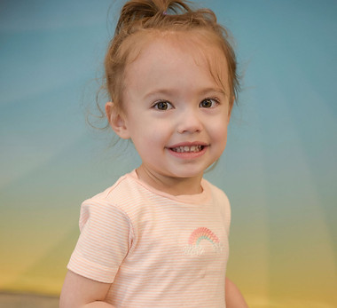 Smiling toddler girl with light brown hair tied up, wearing a light pink striped shirt with a small rainbow design, standing against a blue and yellow gradient background.