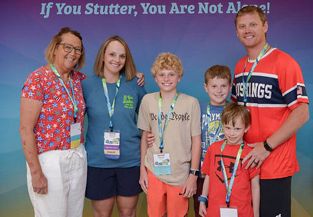 A smiling multigenerational family poses together under a balloon arch at an NSA event, in front of a banner that reads 'If You Stutter, You Are Not Alone!