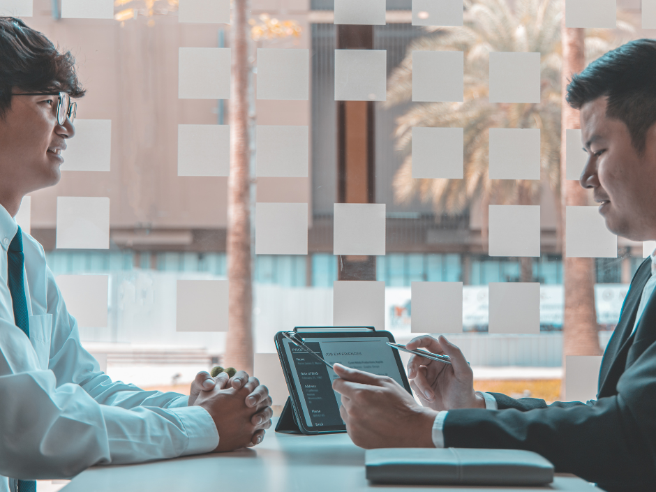 Two men in suits discuss over a tablet showing graphs and text, sitting at a table by a window with frosted squares, palm trees outside.