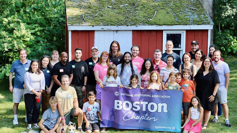 Group of smiling people and a dog pose in front of a rustic shed, holding a "Boston Chapters" banner, set in a lush green garden.