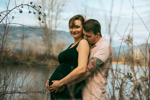 couple poses together showing off pregnant belly, as partner kisses spouses shoulder tenderly.