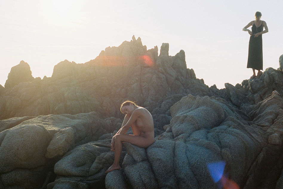 nude woman posing on turquese rocks in Corsica while another woman in a black dress rewinds her analog film to shoot more 