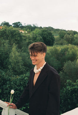 laughing and smiling young man wearing blazer and a shell necklace and holding a white tulip
