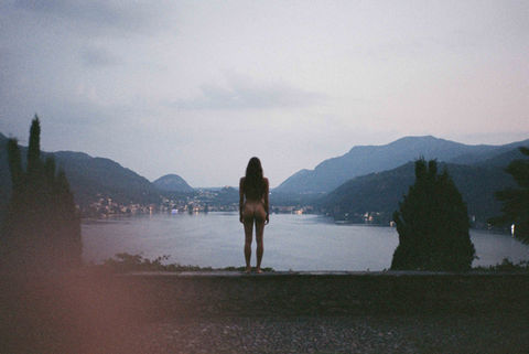 nude woman standing facing a beautiful lake view in evening light