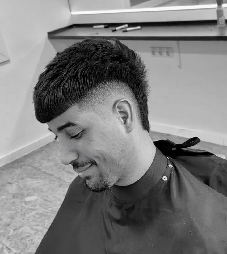 Man with a clean haircut smiles, sitting in a barber chair. Background shows a mirror and shelf with grooming tools. Black and white image.