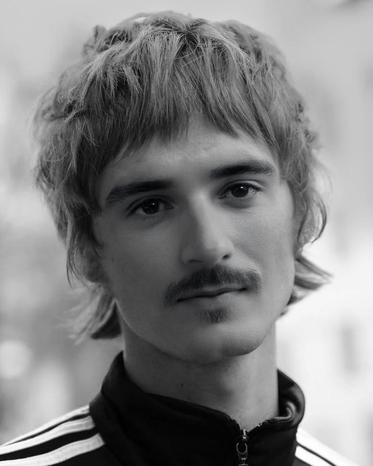 Young man with a mustache gazes forward, wearing a striped jacket. Black and white photo with blurred background, conveying a calm mood.