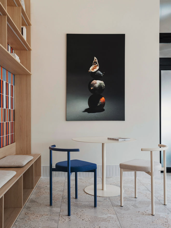 Round white table between a blue upholstered chair and a light beige chair, built-in wooden shelving on the left, and a dark still-life artwork of stacked fruit centered on a white wall.