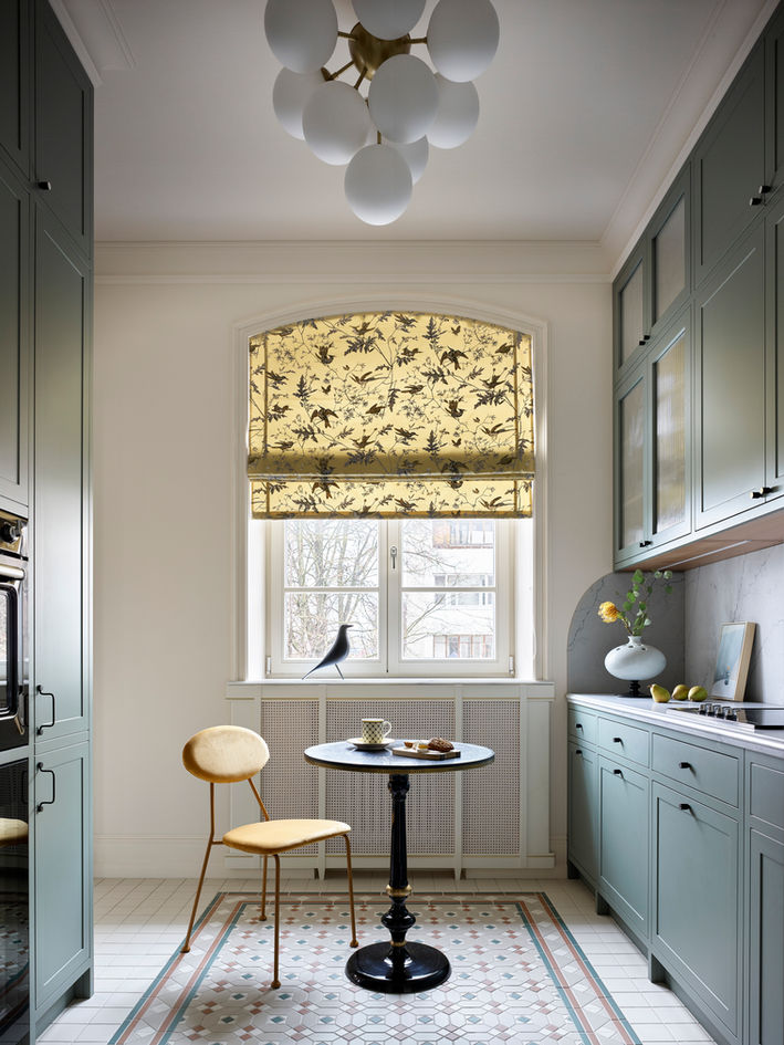 Bright, elegant kitchen nook with sage-green cabinets, patterned tile floor, small round table and chair by a window with a floral Roman shade.