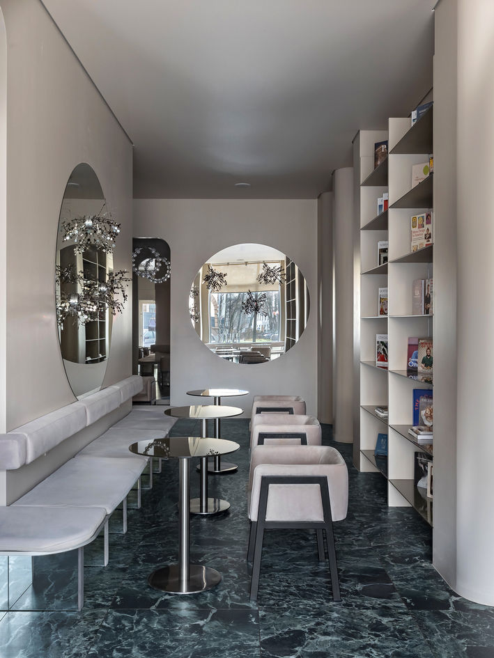Minimalist cafe with curved walls, circular window openings, mirrored wall decor, pale upholstered seating, chrome round tables, tall built-in bookshelves, and a dark marble floor in a softly lit contemporary interior.