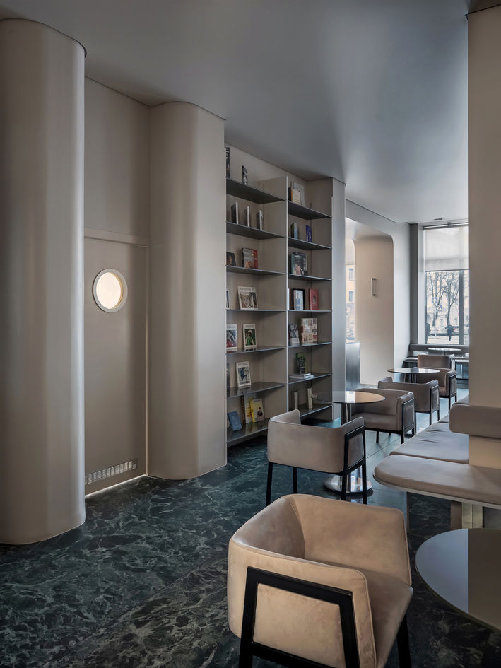 Cafe with curved cream walls, built-in bookshelf filled with books, marble floor, and soft upholstered chairs around small round tables beside a large window.