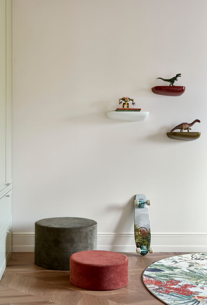 Minimal kids’ room with floating shelves holding dinosaur toys, a skateboard leaning against the wall, and two round floor cushions on a wooden floor.