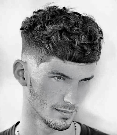 A man with a stylish curly textured haircut and a neatly trimmed beard, showcasing a modern and trendy look against a plain backdrop.