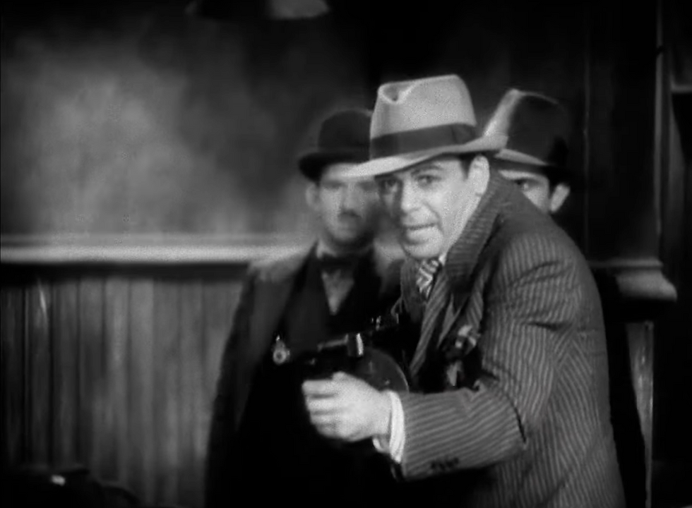 Men in hats and suits in a vintage, smoky room; one holds a tommy gun. The scene is tense and dramatic, evoking a noir film mood.