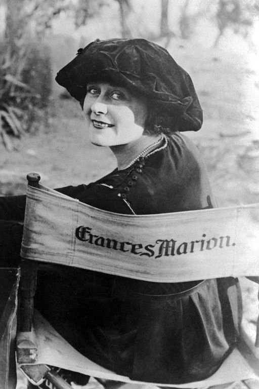 A woman in a black hat smiles at the camera, seated on a director's chair labeled "Frances Marion." The setting appears outdoors, vintage style.