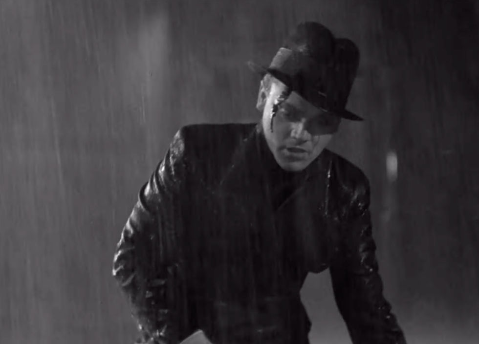 Man in black suit and hat stands in the rain, appearing somber. Monochrome setting with dramatic shadows, creating a noir film mood.