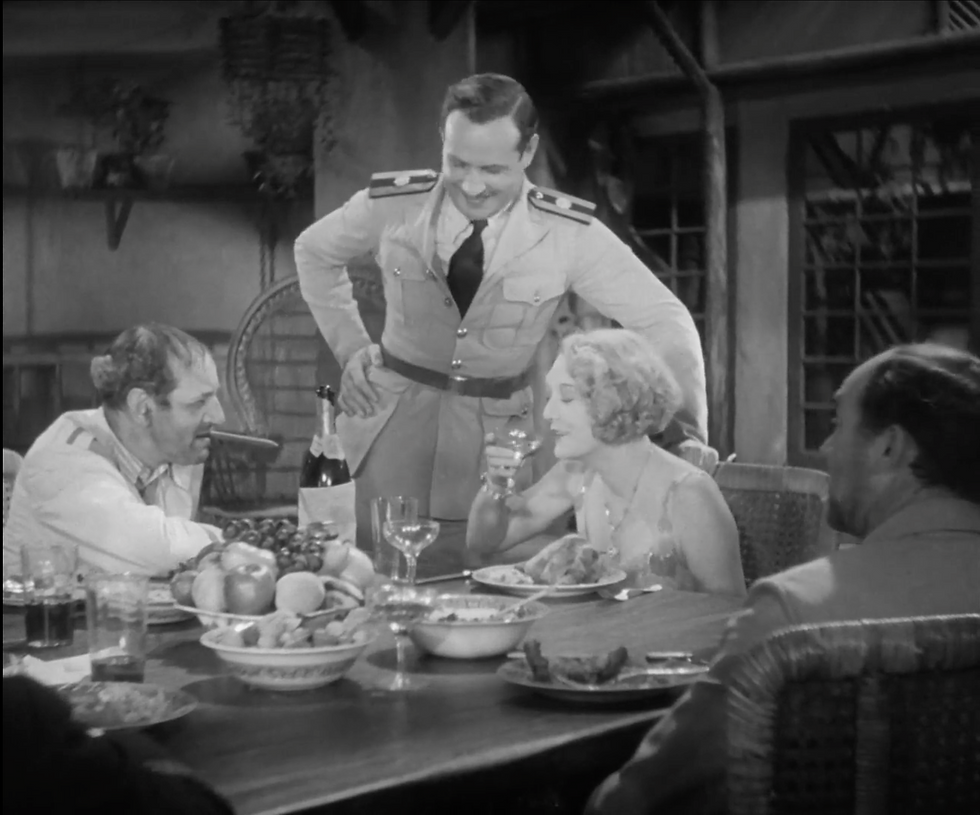 Dining scene with four people; a man in uniform smiles, a woman holds a wine glass. Table filled with food and drink. Cozy setting.